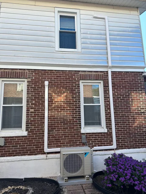 Daikin outdoor mini-split unit on supports with white line-set covers along a brick and sided exterior.